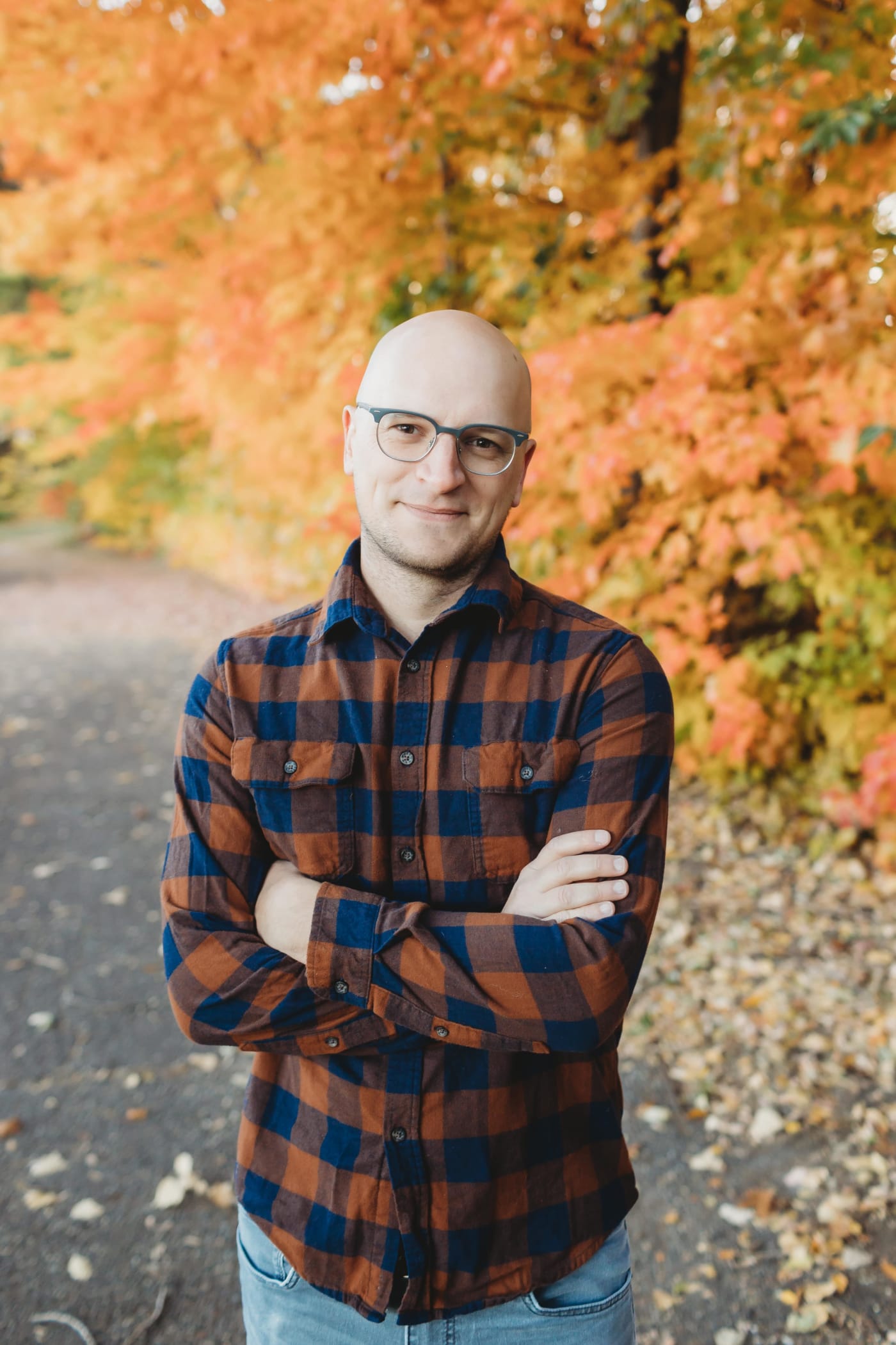 A portrait of Brian Muenzenmeyer. Brian is a 30-somthing bald white man wearing a plaid shirt, grey glasses, and a five o'lock shadow. Photo taken by Samantha Brinda Photography.